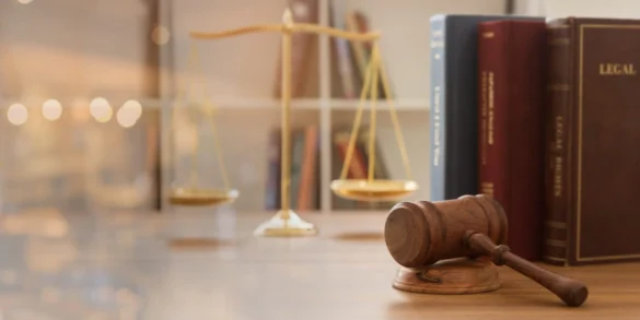 Imagem de cunho advocatício, ilustra, sobre uma mesa, livros sobre leis, um martelo de juíz e uma balança, que representa o símbolo da justiça.