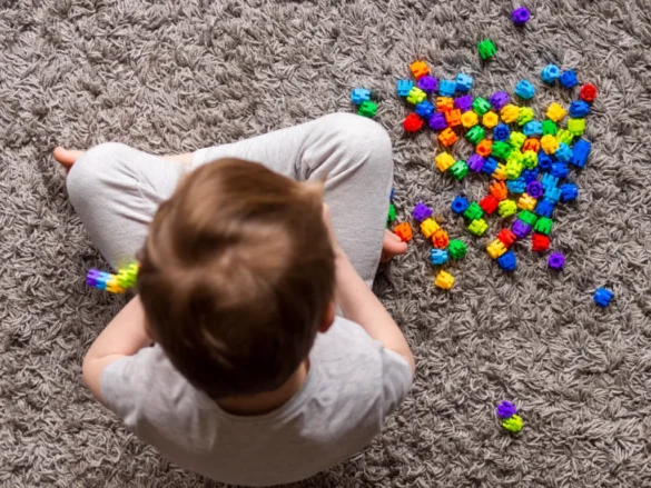 Criança com deficiência intelectual sentada sobre um tapete e brincando com pecinhas coloridas.